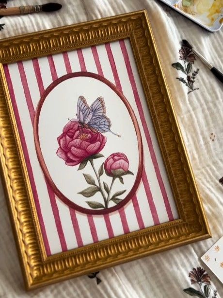 Gold framed watercolor painting featuring a butterfly on a pink peony against a striped background.