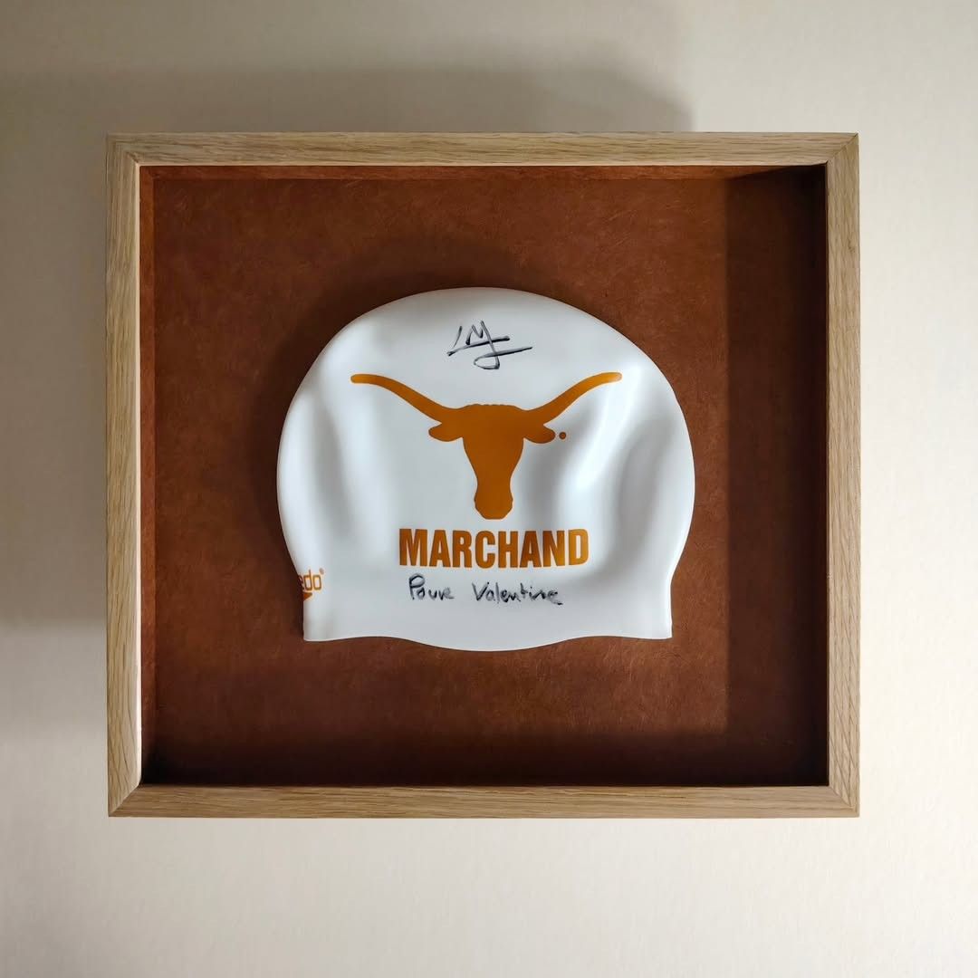 White swimming cap with orange Longhorn logo, 'MARCHAND,' and 'BOB HASTINGS' in a display frame.