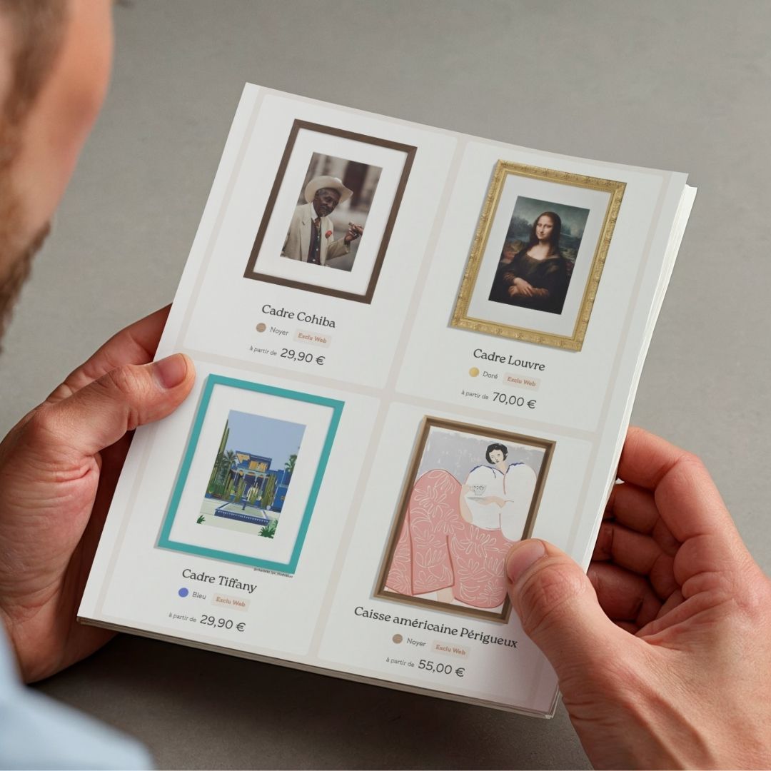 A person's hands holding an open brochure displaying four framed artworks with details.