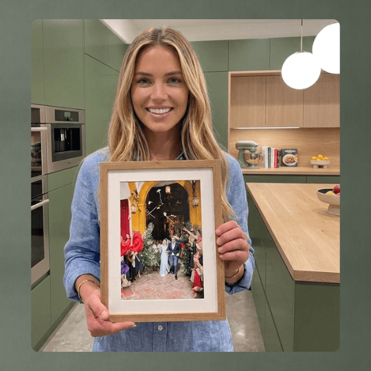A smiling woman in a blue shirt holds a framed photo of a wedding scene.
