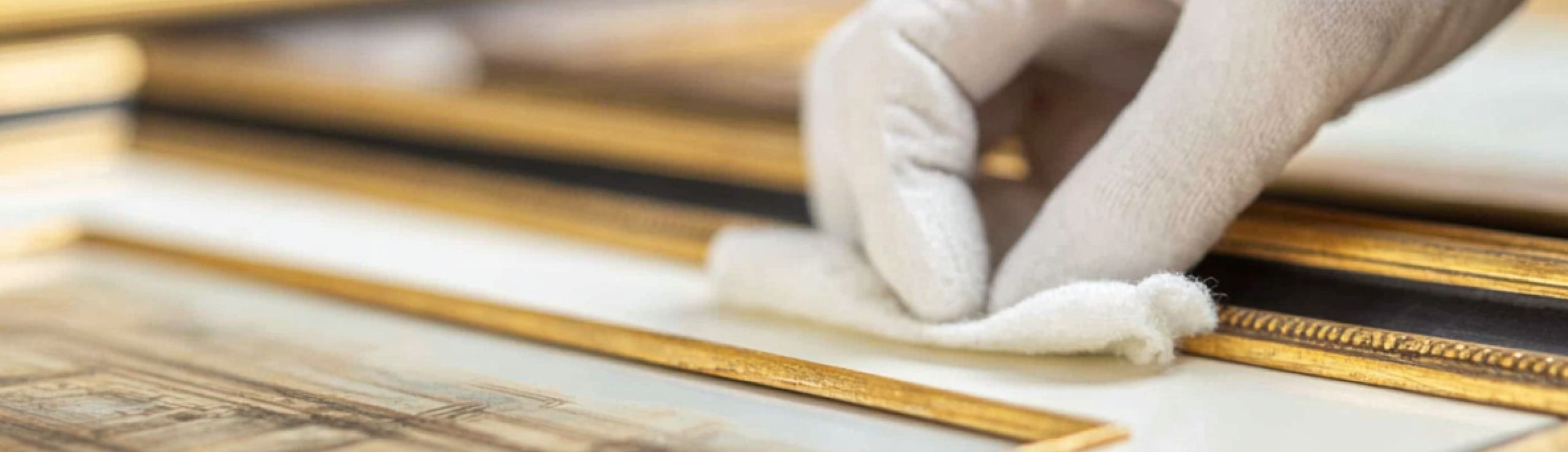 Gloved hand meticulously cleans a golden picture frame with a soft white cloth.