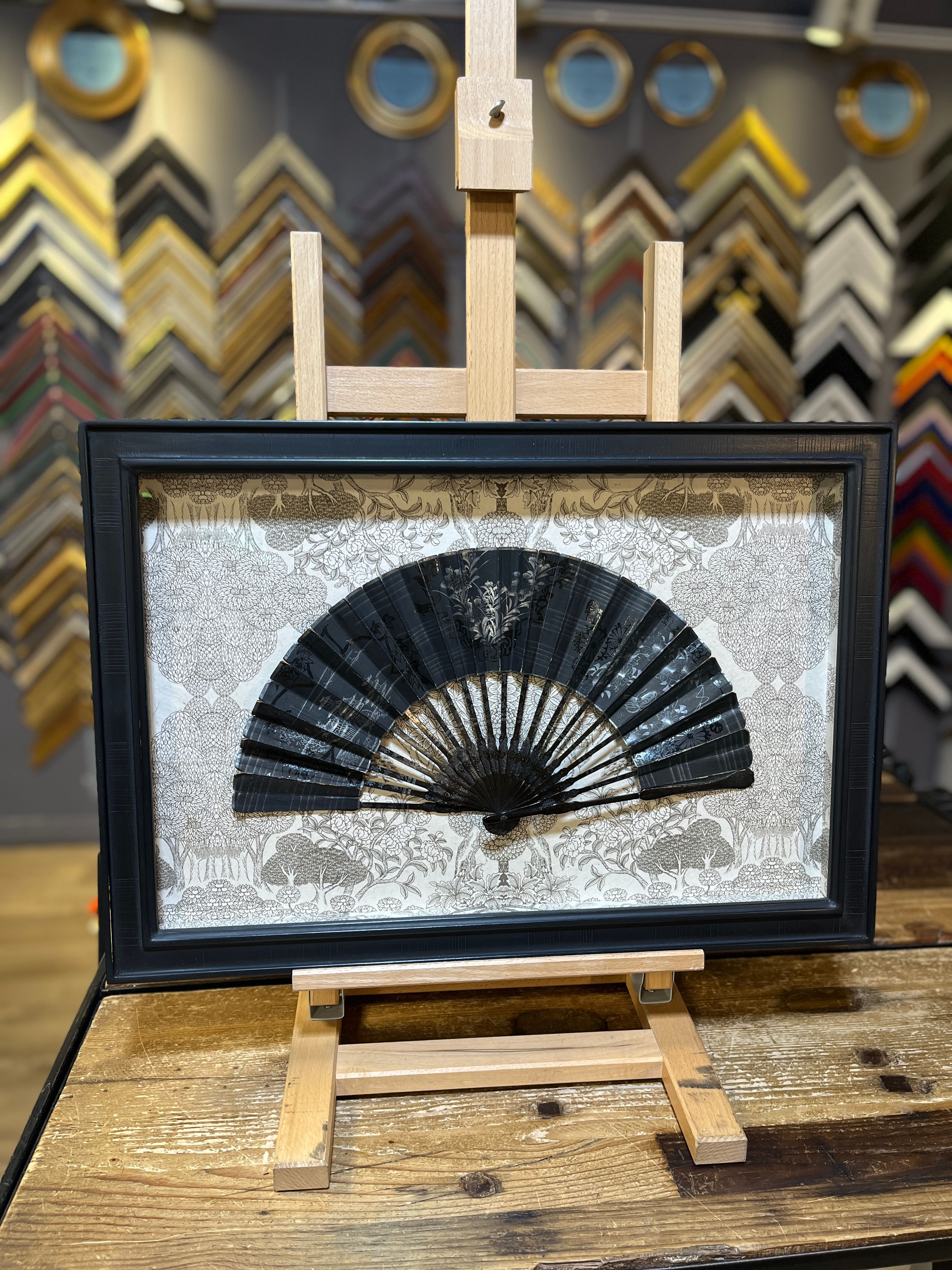 Intricately framed black fan displayed on a wooden easel in a shop setting.