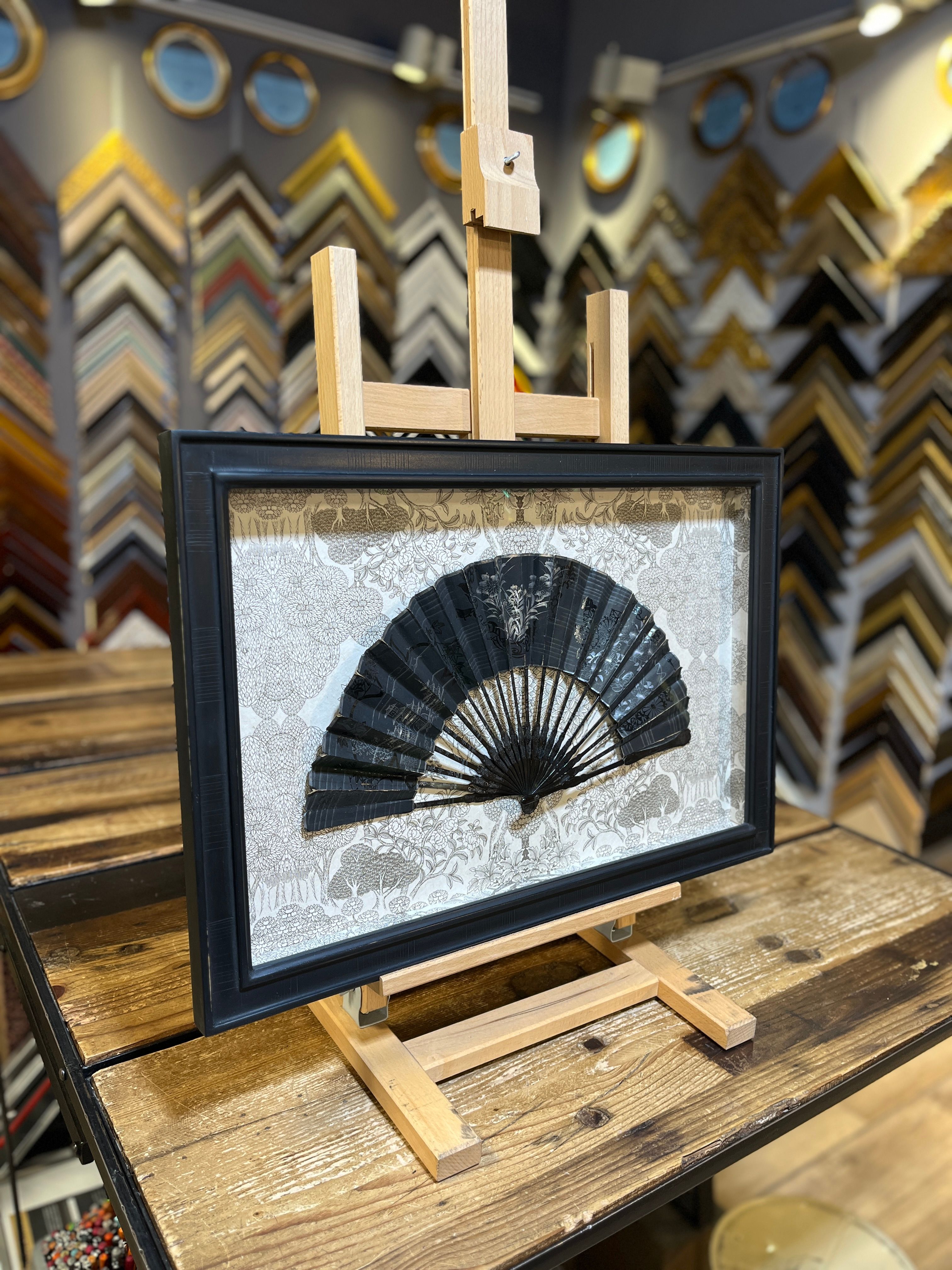 Framed black decorative fan with intricate design displayed on a wooden easel.
