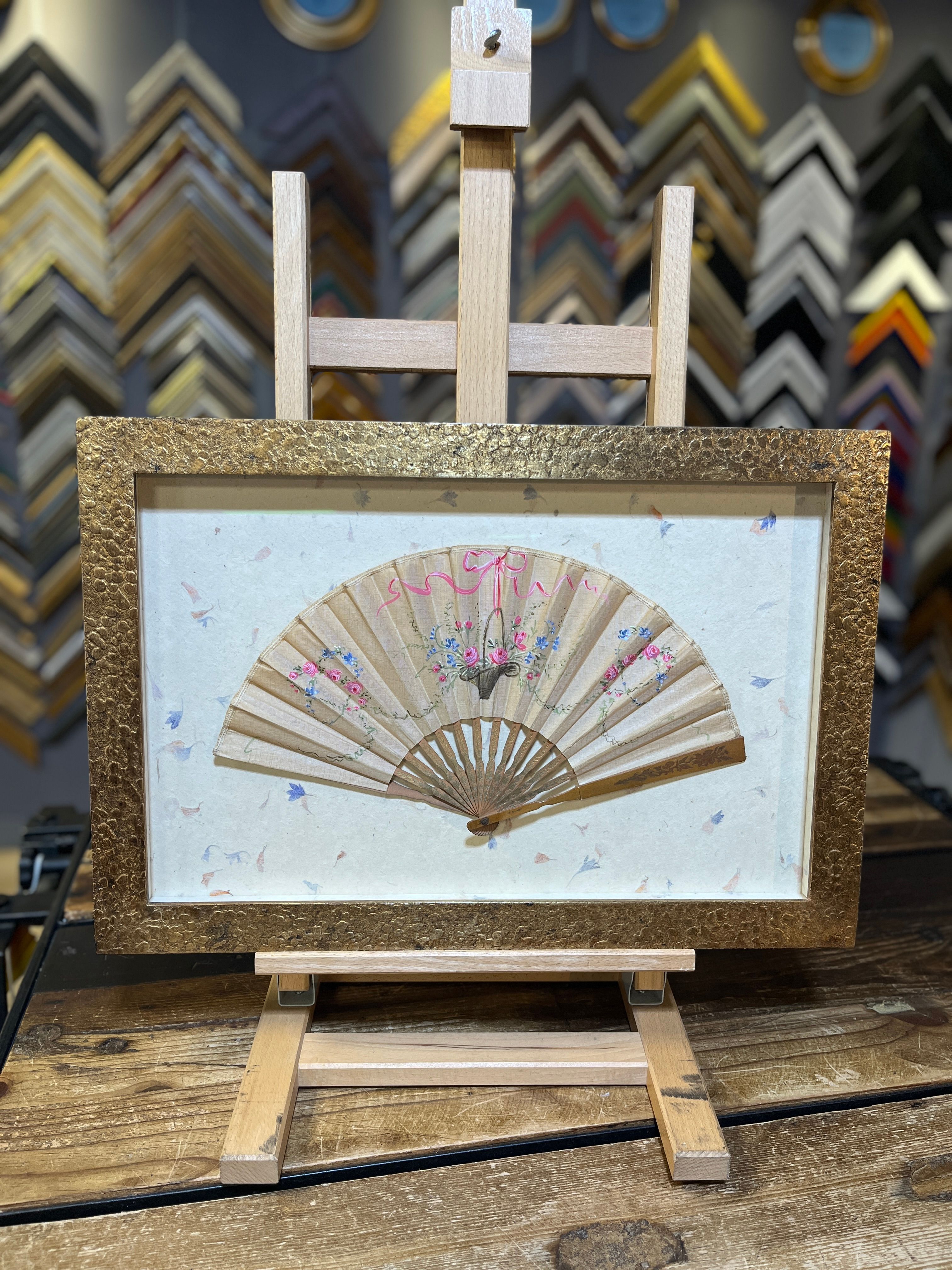 A delicate Japanese fan, open and framed in gold, sits on a wooden easel.