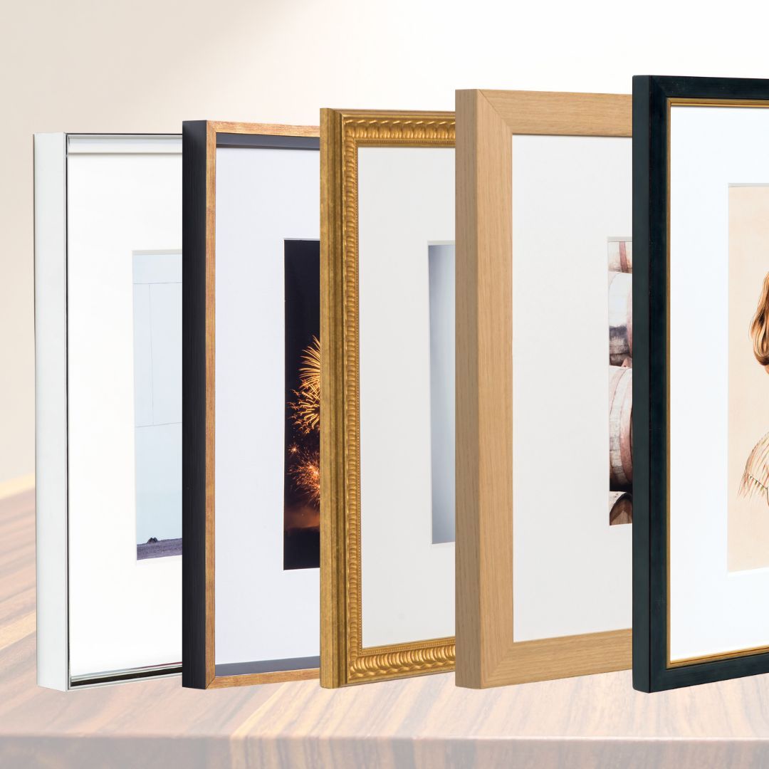 A row of five empty picture frames in white, black, gold, light wood, and dark wood finishes.