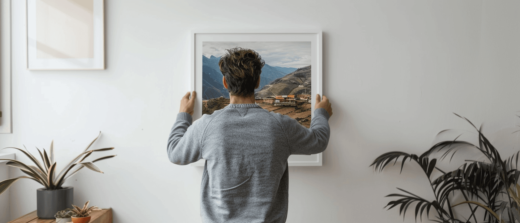 Person hanging a framed mountain landscape picture on a white wall in a modern room.